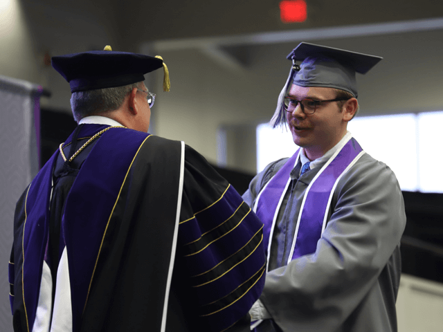 male student in gray graduation gown shakes president's hand and receives diploma on graduation stage